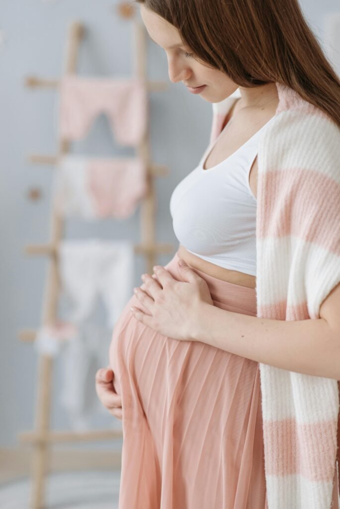 Side view of a pregnant woman indoors, gently holding her belly, wearing a white top and pink skirt.